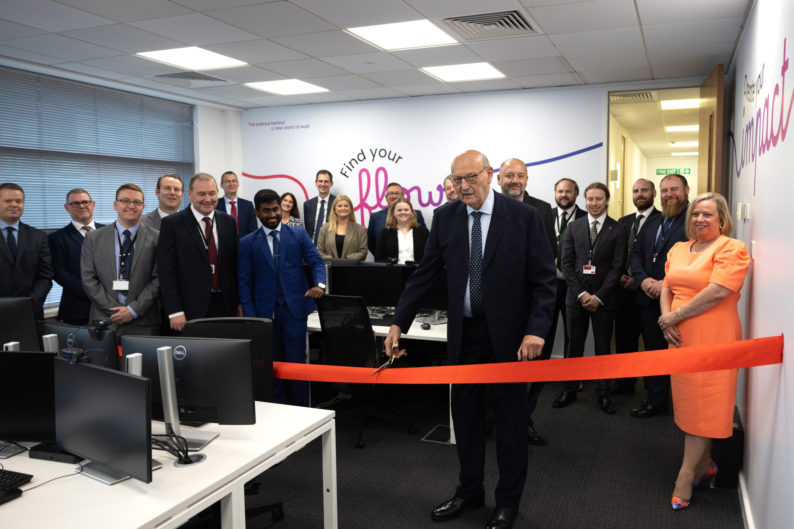 Small office room full of people and a red ribbon in the foreground being cut by a person in a suit.