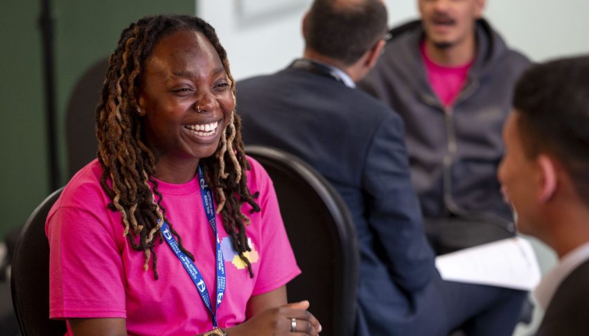 Young people smiling and talking together in a room wearing lanyards and holding sheets of paper.