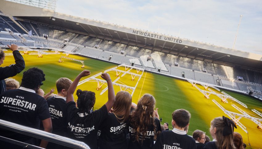 Children in the stands of St James' Park wearing navy Adidas t-shirts made specifically for the Mercury Prize Fringe.